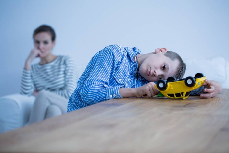 Image of boy slumping on a table with an upside down toy car in his hands as a concerned parent looks on from behind him, out of focus