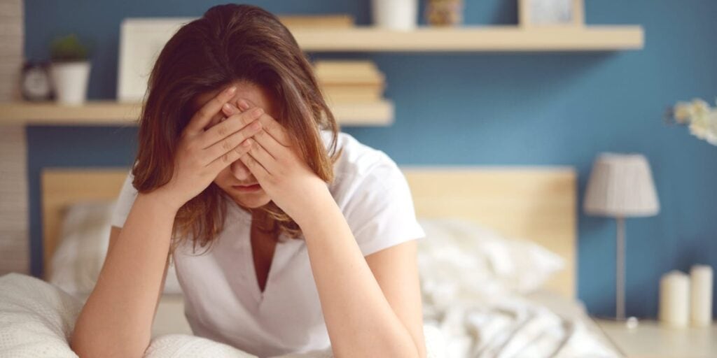 Woman sitting up in bed, with her head in her hands, obscuring her face, but indicating headache or general suffering